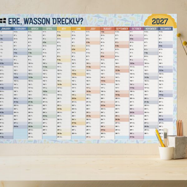 Large wall-planner fixed to a wall by a desk. The wall-planner features a Cornish pattern and reads "Ere, wasson dreckly?". The desk contains mixed stationery, vase & mobile phone